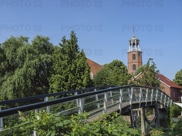 Wooden bridge leading to a church tower, surrounded by lush green vegetation and blue sky, Ditzum,