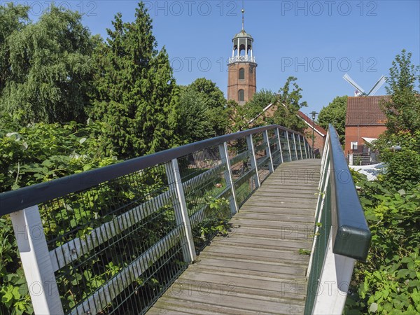 Wooden bridge leads to a church tower and a windmill, surrounded by trees under a blue sky, Ditzum,