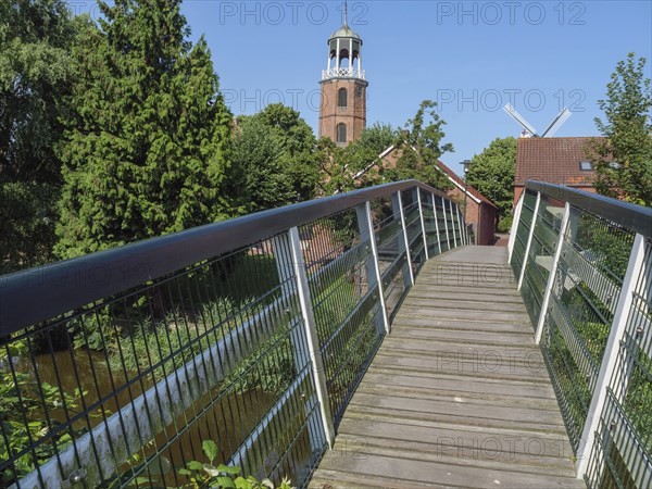Wooden footbridge leading to a historic church tower, surrounded by green vegetation and clear