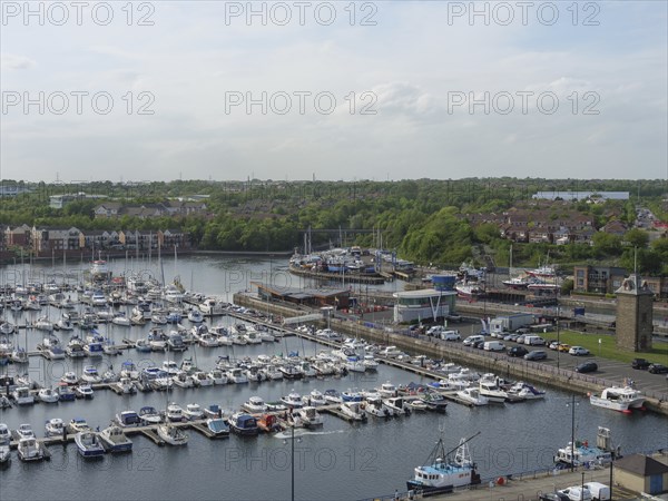 A harbour full of boats and yachts, surrounded by green spaces and houses under a cloudy sky,