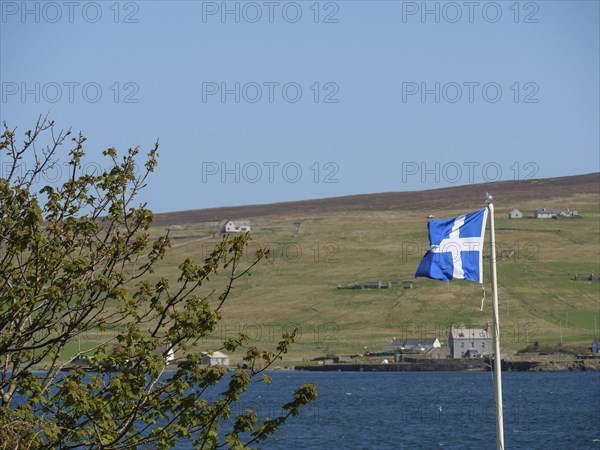 A Scottish flag flies on the coast with green hills and scattered houses in the background,