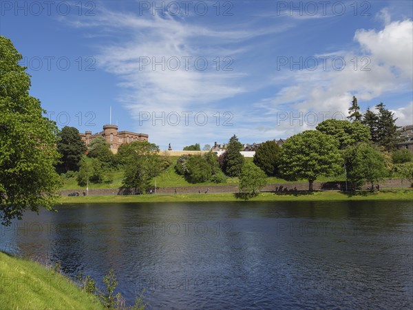 A river flows in front of a green hill and a castle, surrounded by lush vegetation and a clear sky,
