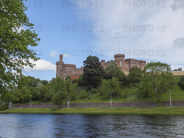 A castle overlooking a river, surrounded by green banks and trees under a blue sky, inverness,