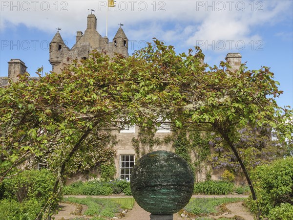 Castle with towers in the background, an archway of plants and a sculpture in a well-tended garden,