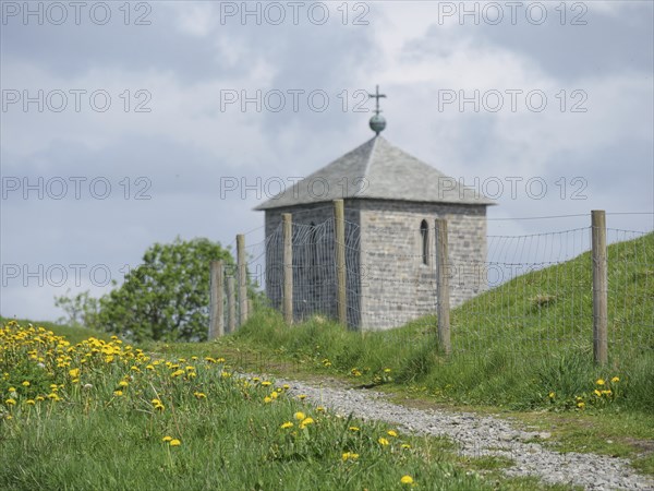 Stone church with picket fence on a green path, surrounded by yellow flowers and cloudy sky,