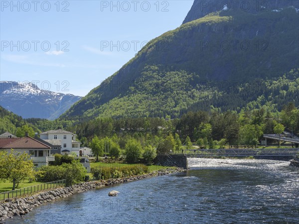 A river with a bridge, flanked by houses and trees, embedded in a green valley with mountains in