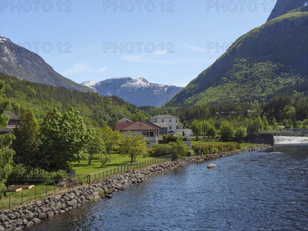 A river flows past houses and trees in a green valley surrounded by high mountains, Eidfjörd,
