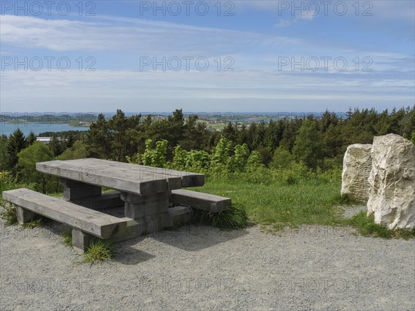 Wooden table and bench on a gravel path with a view of the forest and sea under a slightly cloudy