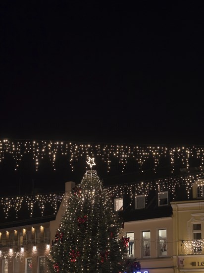 Large Christmas tree with festive lights and star on top, surrounded by illuminated buildings at
