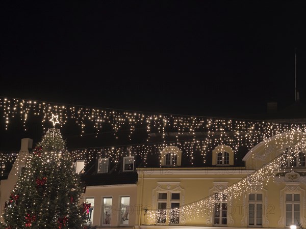 Festively illuminated Christmas tree and buildings on a dark night. Fairy lights hang over the