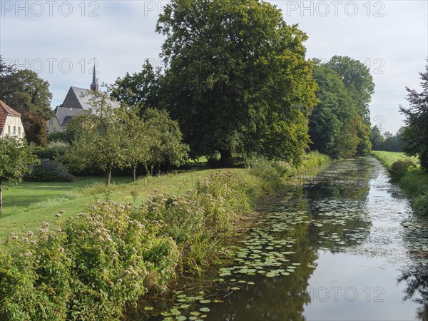 River with tree-lined bank, meadows and church tower in the background in calm weather, marienthal,