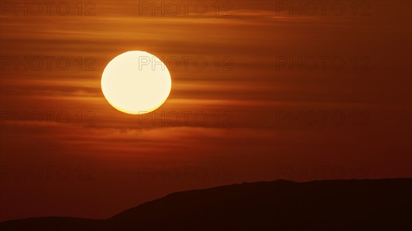 A large, bright ball of sunlight in the sky sinks behind the dark silhouettes of hills, offshore