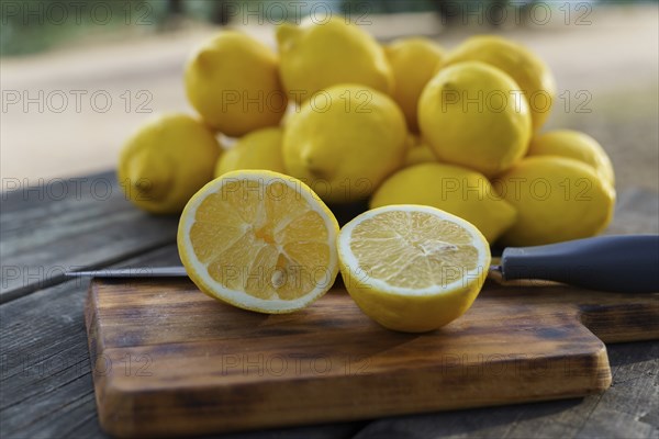 Lemon cut with a knife on a wooden board on a table in a park with a pile of lemons and trees in