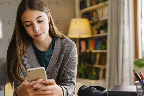 A girl in a grey jumper sits in front of a bookshelf and concentrates on using her smartphone, AI