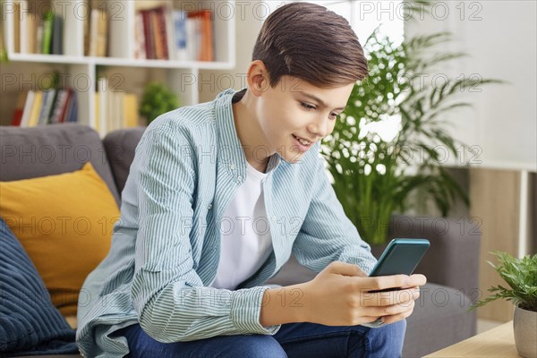 A smiling boy in a striped shirt sits on a sofa and uses his smartphone, AI generated, AI generated