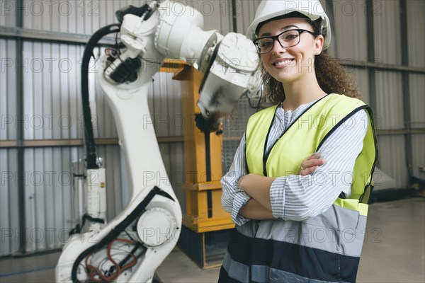 Woman with helmet and yellow waistcoat in a factory next to a large white robot arm, surrounded by