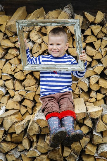 A smiling boy in a striped shirt and boots sits on a woodpile, holding a rustic, weathered frame