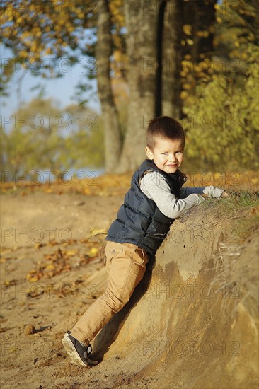 A young boy smiling and climbing a dirt slope in an autumn forest setting with colorful fall