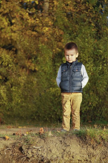 A young boy in a blue vest stands on a grassy area surrounded by autumn leaves and trees, Belarus