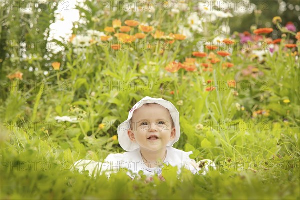 A smiling baby wearing a white hat sits amidst a garden filled with colorful flowers and lush