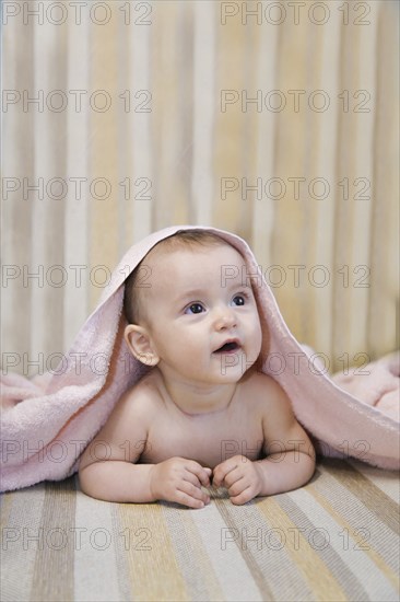 A baby lying on a striped surface, partially covered with a pink towel, looking up with an adorable