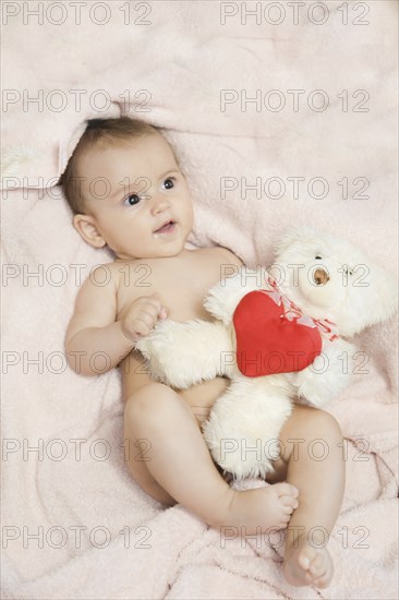 A baby lying on a pink blanket holding a white teddy bear with a red heart