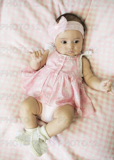 Baby in a pink dress with a headband and bow, lying down on checkered bedding, wearing white shoes