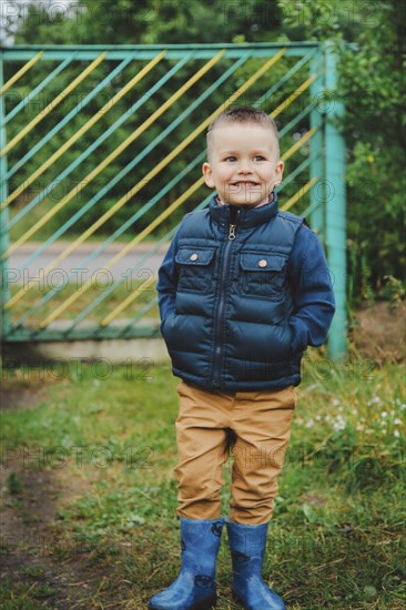 Young boy wearing a blue jacket and boots stands outdoors near a green gate.Belarus.Minsk