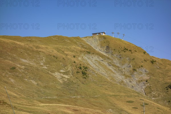 Extensive mountain landscape with a hut on a hill, crossed by a cable car under a blue sky,