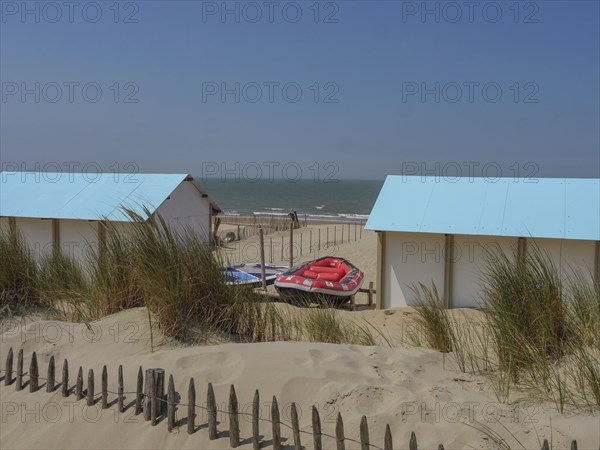 Wooden huts on the beach, surrounded by sand dunes and lifeboats, with the sea in the background,