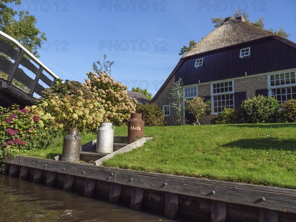 A house with thatched roof and well-tended garden, flowers and milk cans in the foreground, bright