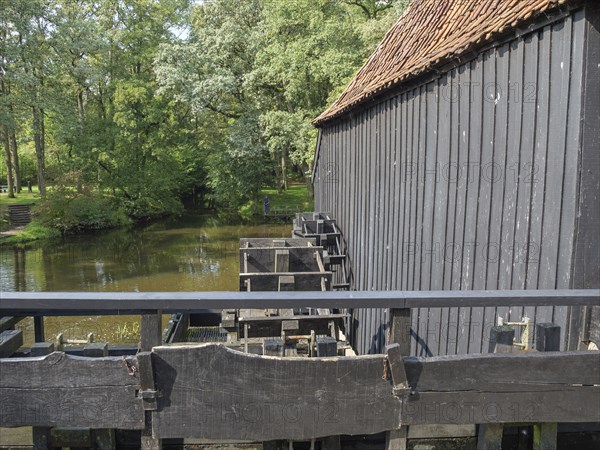 A historic wooden building next to a river and a bridge, surrounded by dense forest, haaksbergen, .