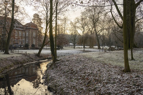 A small stream with a reflection flows through a frosty park, in the background a large building