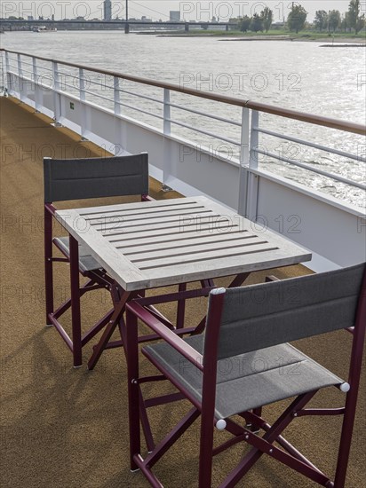 Deck of a ship with table and chairs, view of the river and the surrounding landscape. The railing