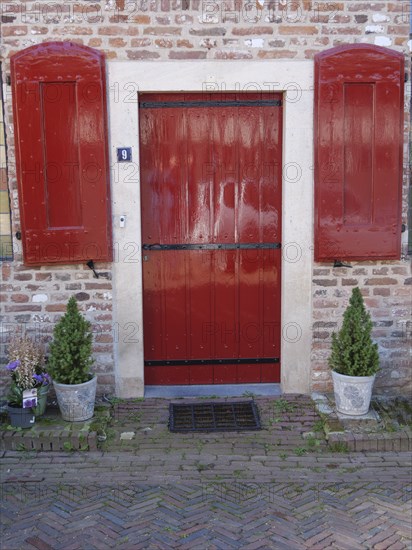 Red door with shutters surrounded by brick wall and potted plants, Doesburg, Netherlands
