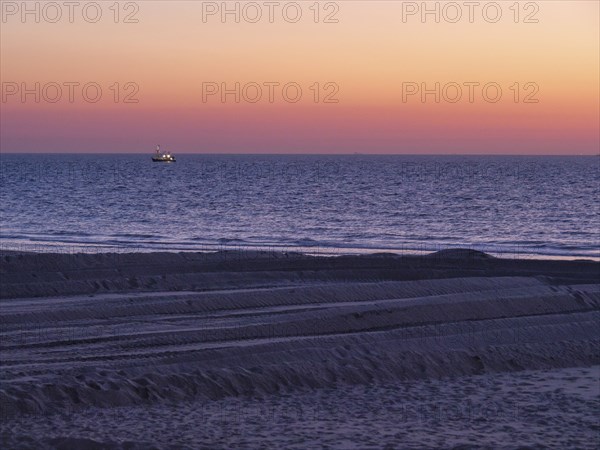 A boat sails on the sea during sunset, the beach shows traces in the sand and the atmosphere is