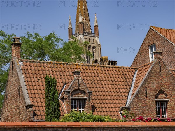 Roofs and a church tower rise behind historic brick buildings under a clear sky, Bruges, Belgium,