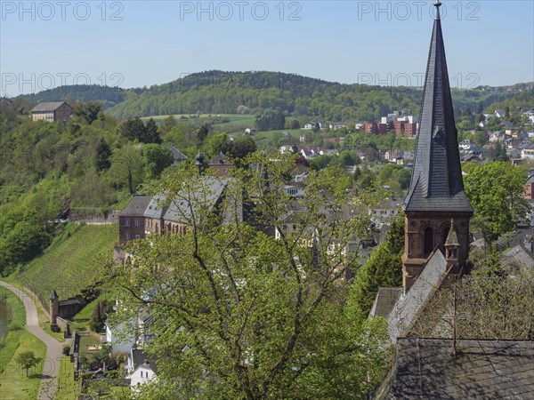 A view over a town with a striking church spire, surrounded by green hills and trees, Saarburg,