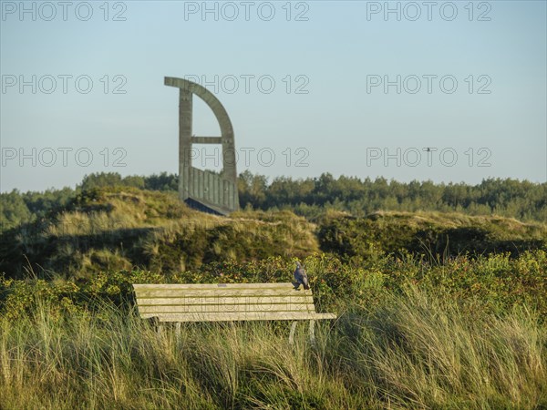 A park bench stands next to a meadow and a sculpture in the background under a clear sky, langeoog,