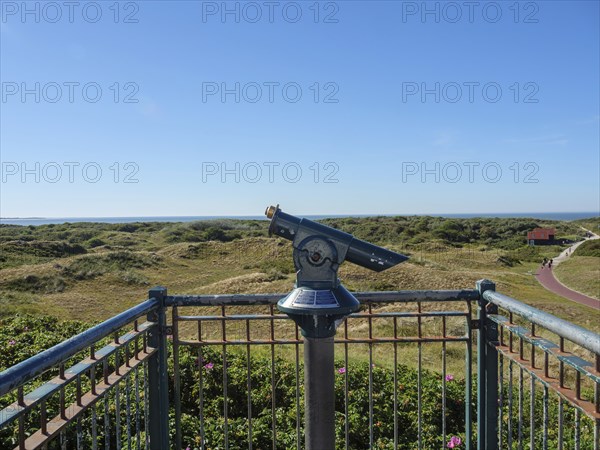 A pair of binoculars on a viewing platform with a view of a vast dune landscape and the sea,