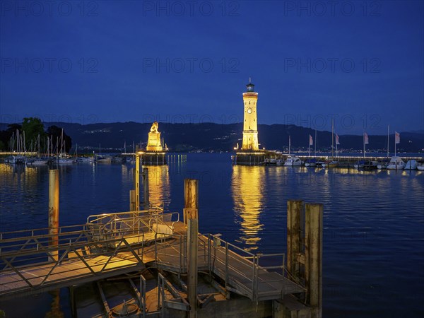 Illuminated harbour at night with lighthouse and lion sculpture reflected in the water, Meersburg,