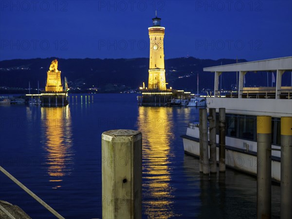 A lighthouse and a statue illuminated at night in the harbour with reflections in the calm water,