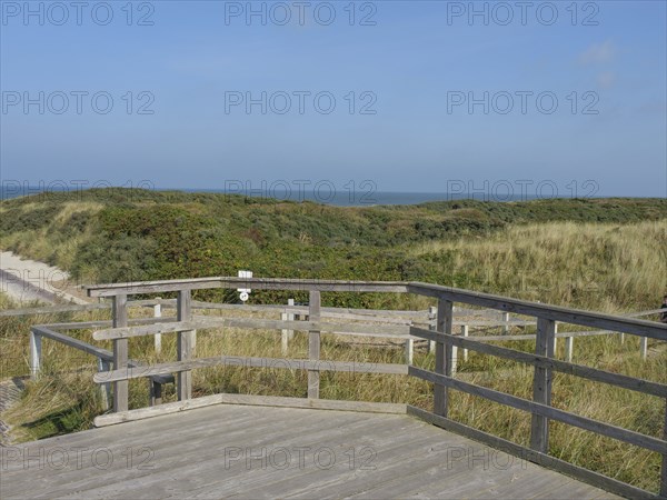 A wooden terrace overlooks the grassy dunes and the calm sea, a peaceful atmosphere dominates the