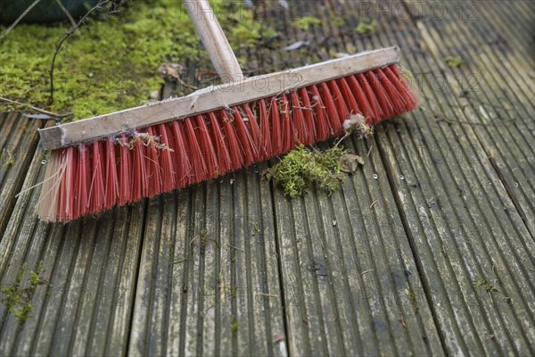Outdoor broom with red plastic bristles on a weathered wooden deck with slippery algae and moss,