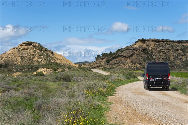 Black van on a gravel road in a hilly, green landscape under a blue sky with clouds, motorhome,