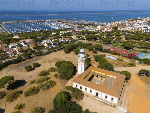 A lighthouse overlooking a coastal town and a marina, surrounded by trees under a sunny sky, aerial