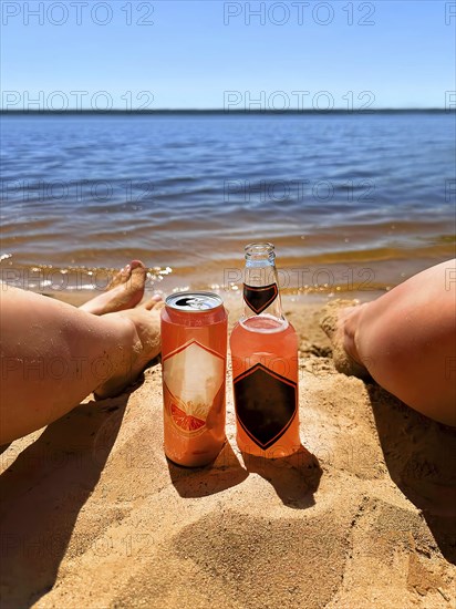 Two people relaxing on a sandy beach with a bottle and can of drink by the water on a sunny day