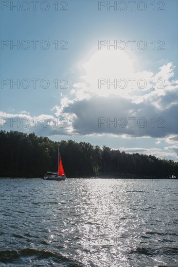 Sailboat with a red sail on a lake under a bright sun and scattered clouds, surrounded by forest