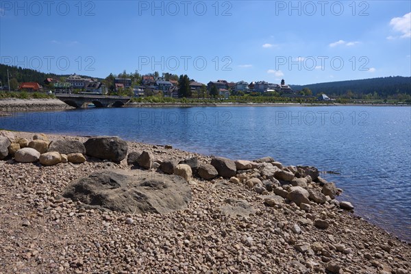 Quiet lake landscape, Schluchsee with pebble beach, bridge and houses on a hill under blue sky,