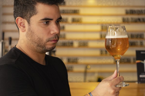 A young Latino man enjoys a craft beer in a pub. He carefully observes the color and hops in the
